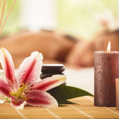 Perfumed sticks, massage oil, stones, fresh flower and scented candles on the table. Woman getting a hot stone massage in the background.